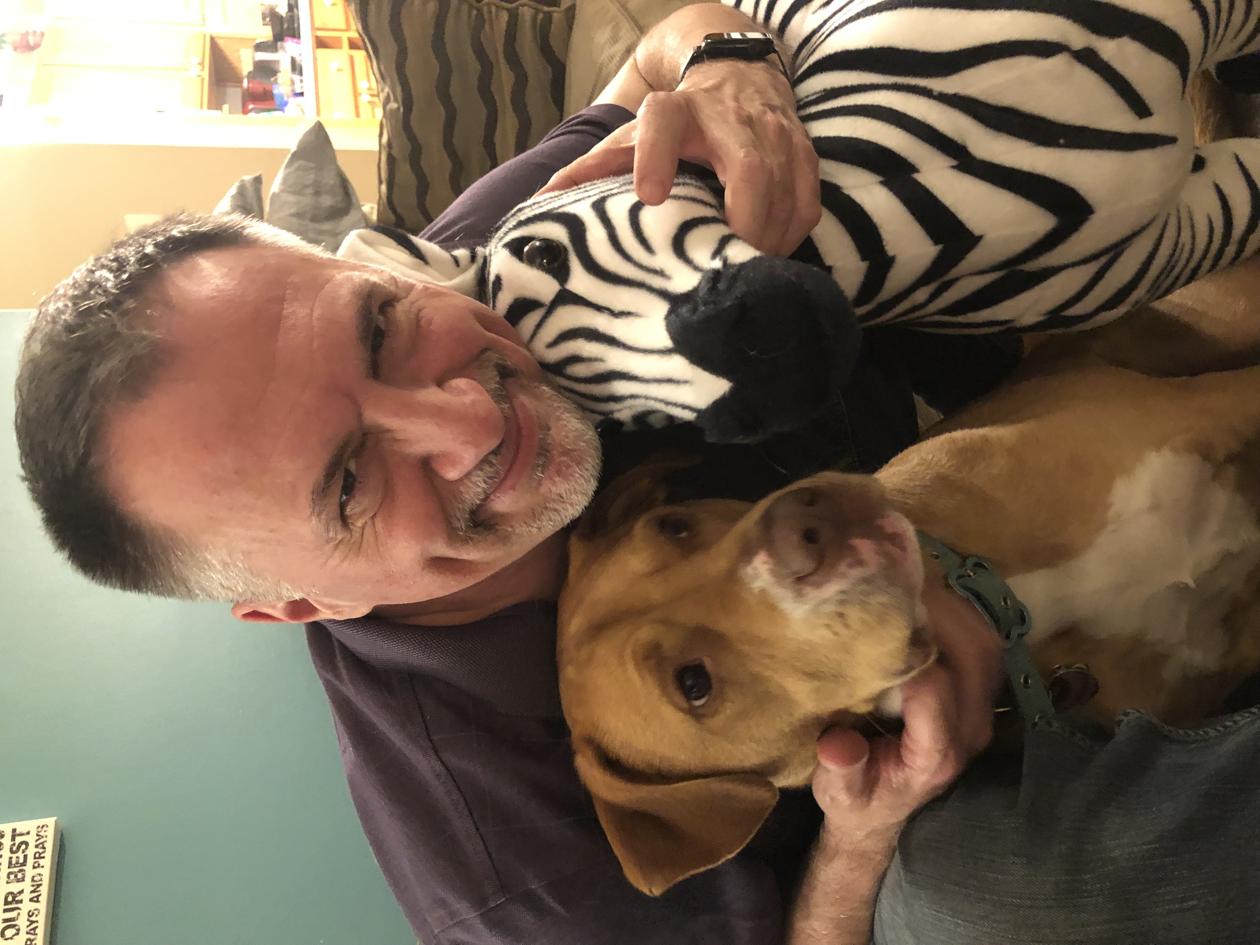 John on a couch hugging Frank the zebra and Tanner the dog simultaneously, all three grinning